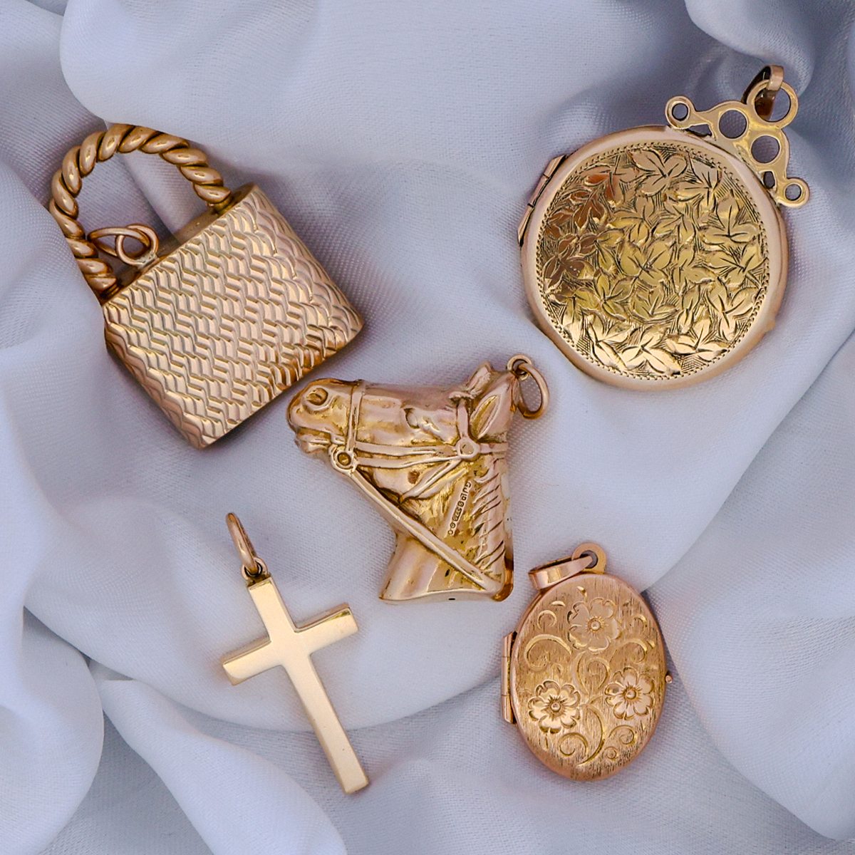 Collection of gold jewellery pendants including a padlock, horse head, cross, oval shaped locket and round shaped locket on a white fabric background.