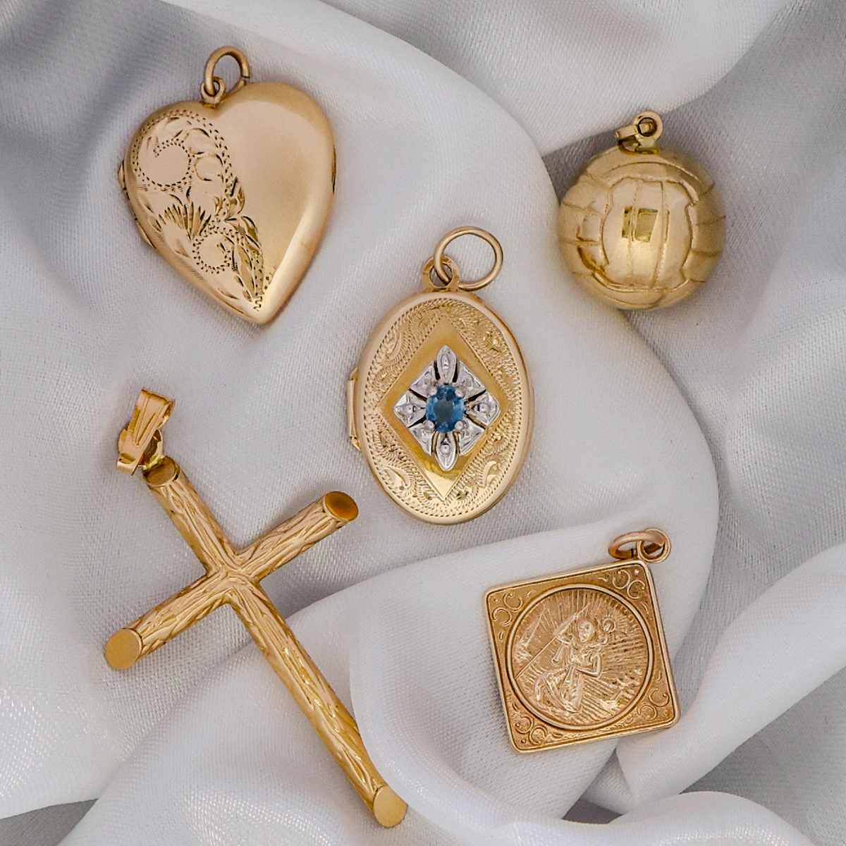 Collection of gold jewellery pendants including a St Christopher, cross, football, heart shaped locket and sapphire set oval shaped locket on a white fabric background.