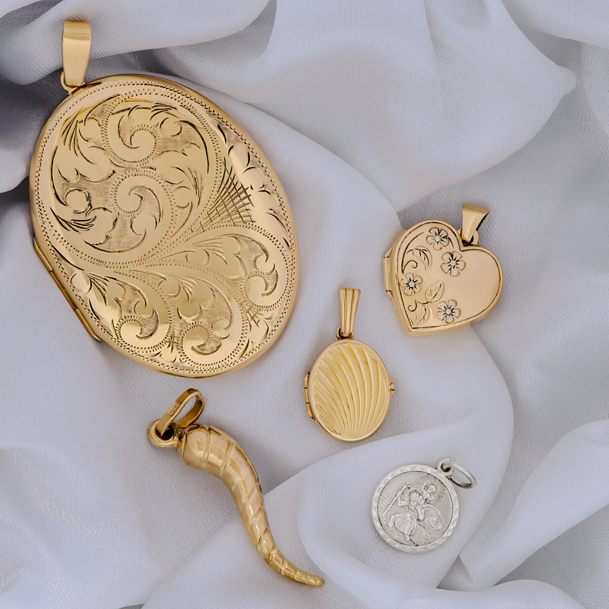 Collection of gold and silver jewellery pendants including a St Christopher, horn of life, heart shaped locket and large oval shaped locket on a white fabric background.